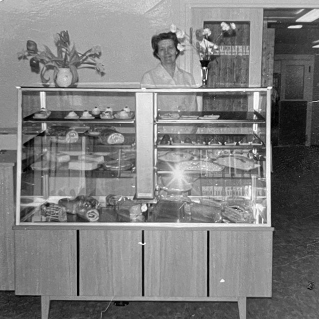 Svart-hvitt foto av en smilende ekspeditør bak en glassdisk med kaker og brød i bakeriutsalget.