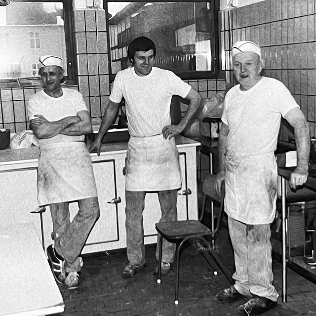Svart-hvitt foto av tre smilende bakere i hvite forklær på bakeriet, to av dem med bakerhatt.