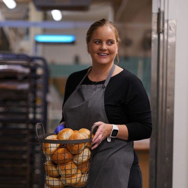 Smilende ansatt i stripete forklær bærer en metallkurv full av nybakte rundstykker gjennom bakeriet.